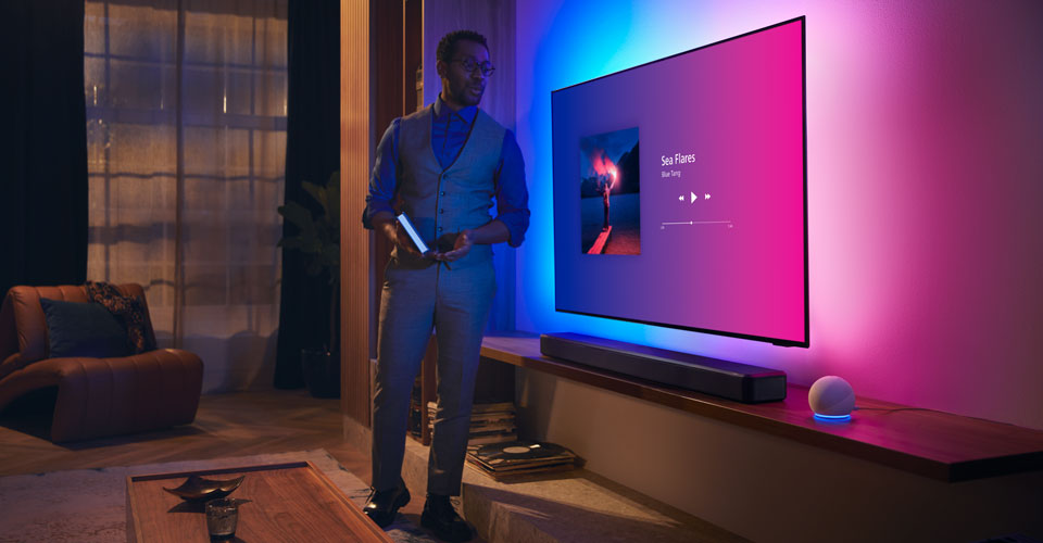 Egy szem&eacute;ly egy modern nappaliban egy Philips Ambilight TV k&ouml;zel&eacute;ben, zenei interf&eacute;sz&eacute;vel &eacute;s Fidelio soundbarral