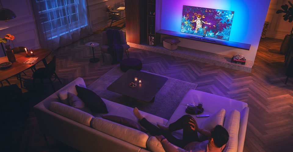 F&eacute;rfi pihen a kanap&eacute;n egy halv&aacute;nyan megvil&aacute;g&iacute;tott nappaliban, &eacute;s Philips Ambilight t&eacute;v&eacute;t n&eacute;z.