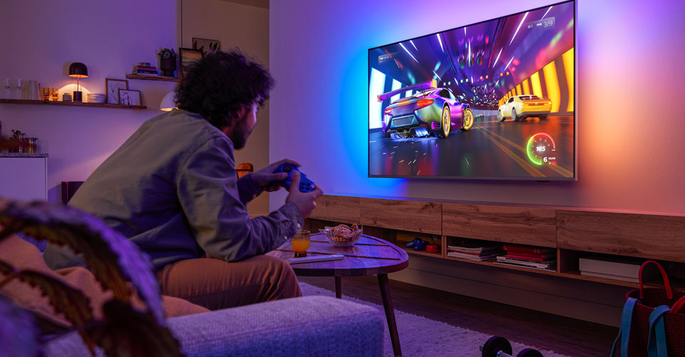 Egy szem&eacute;ly a kanap&eacute;n &uuml;l, &eacute;s egy aut&oacute;versenyzős videoj&aacute;t&eacute;kot j&aacute;tszik egy Philips Ambilight t&eacute;v&eacute;n
