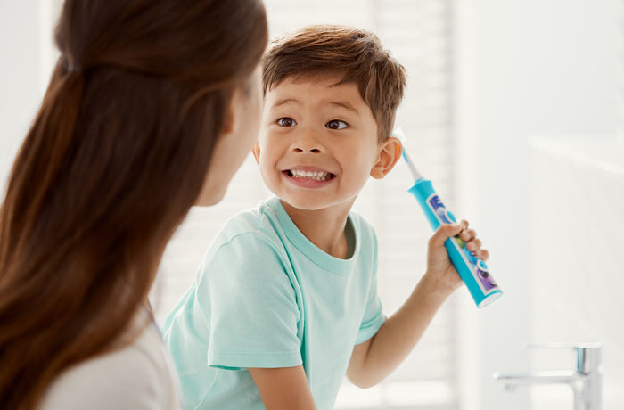 Egy kisfi&uacute; grimaszolva &eacute;desanyja fel&eacute; mutatja fogsor&aacute;t, mik&ouml;zben egy k&eacute;k Philips Sonicare for Kids gyermek fogkef&eacute;t tart a kez&eacute;ben. 