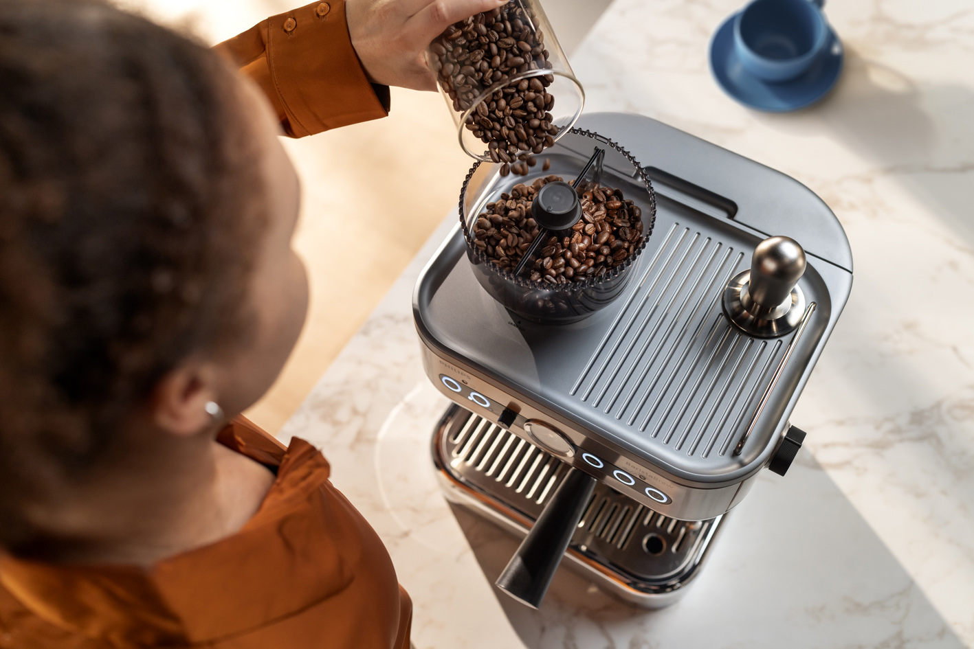 Egy fiatal h&ouml;lgy szemes k&aacute;v&eacute;t t&ouml;lt a Philips Barista Brew k&eacute;sz&uuml;l&eacute;k szemesk&aacute;v&eacute;-tart&aacute;ly&aacute;ba